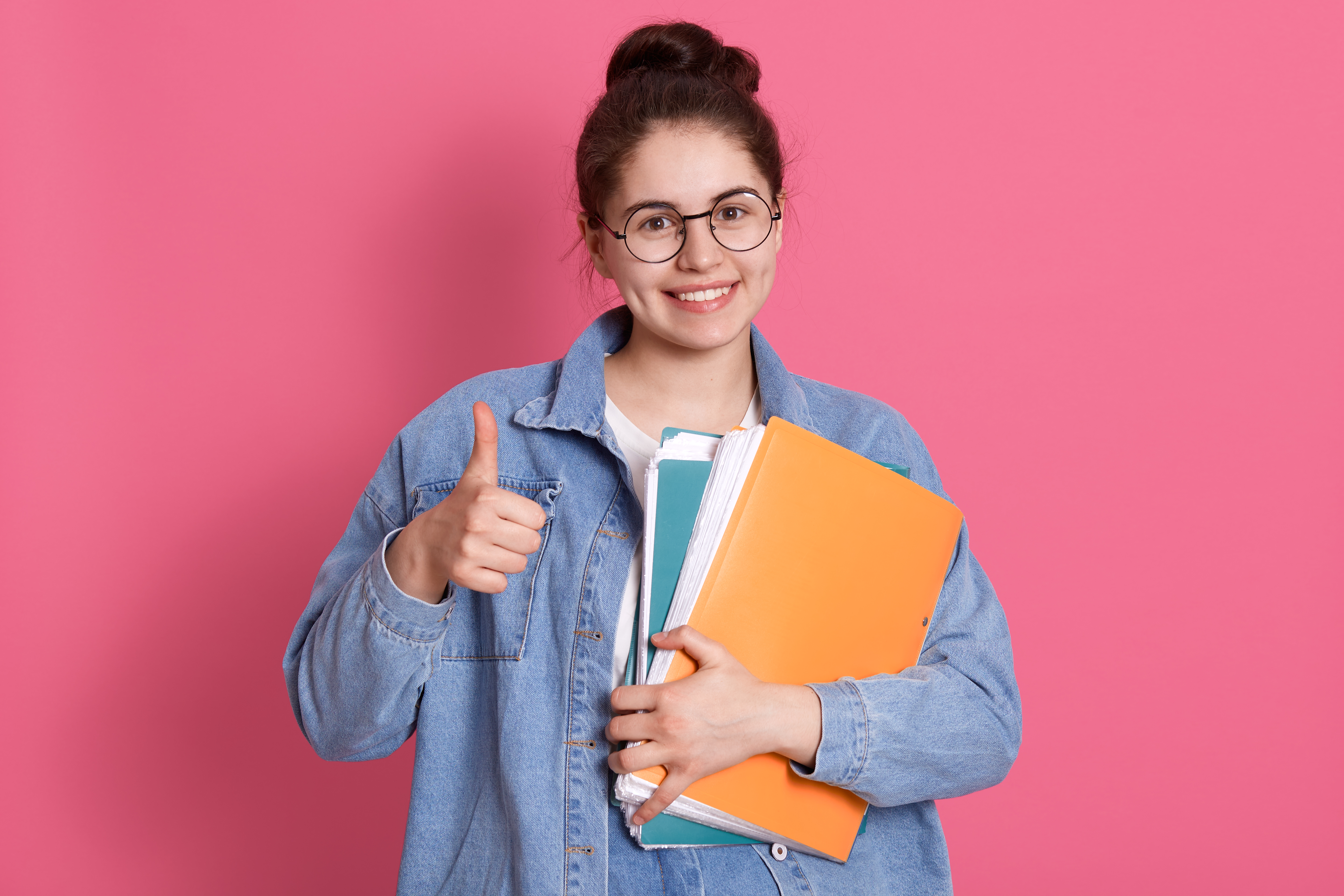 young-student-woman-wearing-denim-jacket-and-eyeglasses-holding-colorful-folders-and-showing-thumb-up-on-pink.jpg